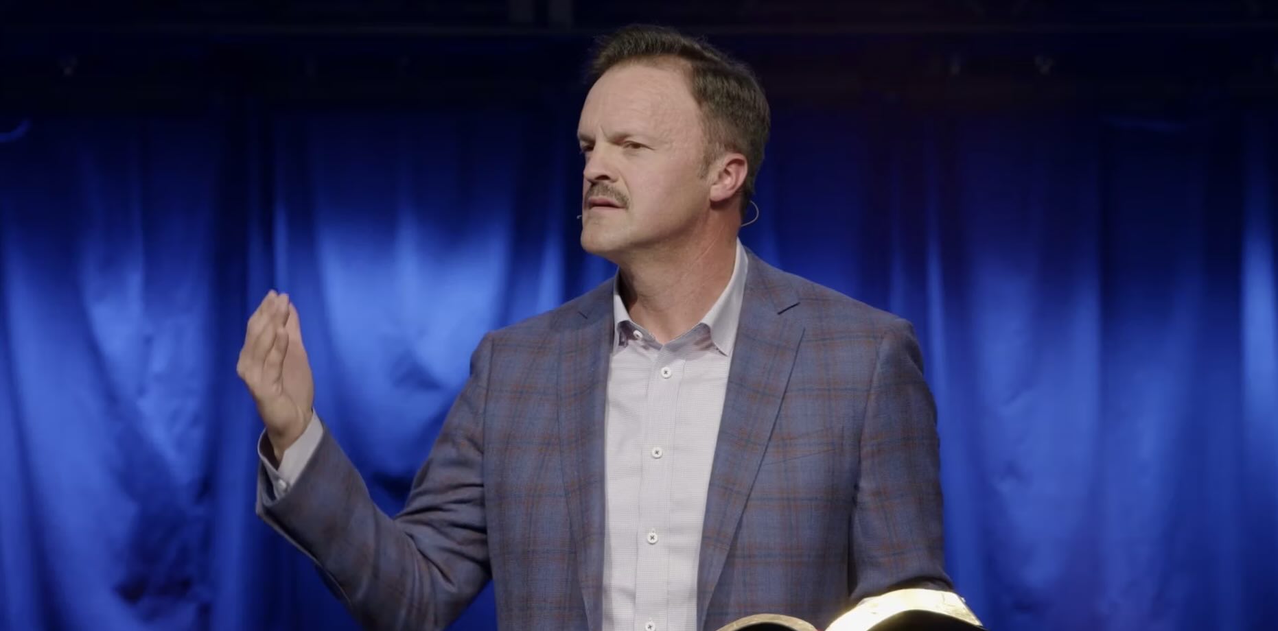 Man with a mustache preaching with a Bible in his hand.