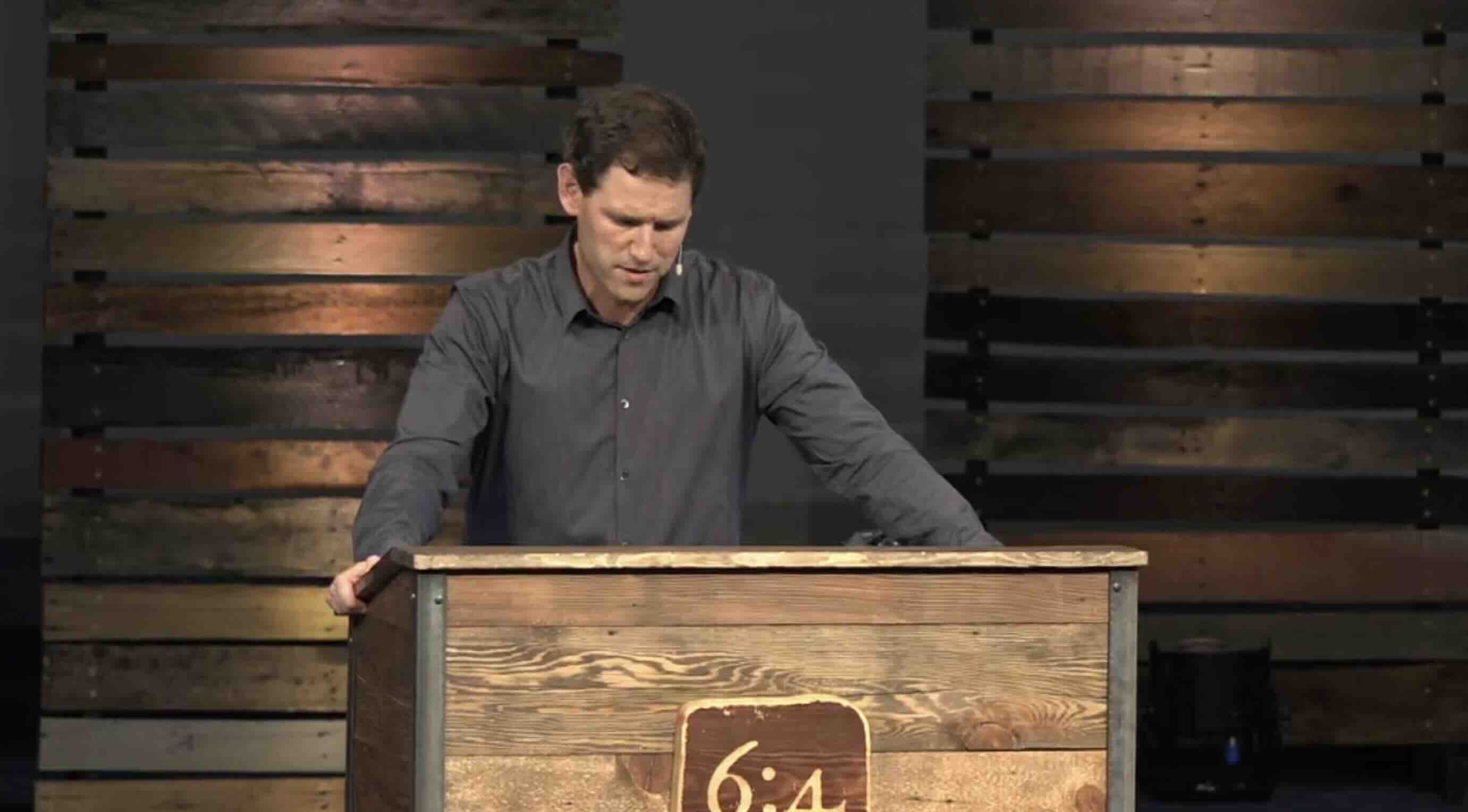 Man in grey long-sleeved shirt standing at a wooden podium with 6:4 on the front